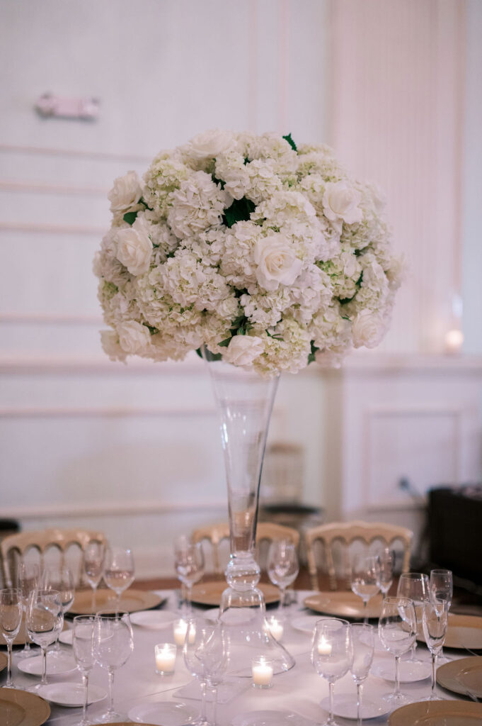 Large centerpiece of white roses and hydrangeas sits on glass pedestal on round wedding guest table | Wedding Photography by Lauren Bliss at the Cescaphe Ballroom Philadelphia