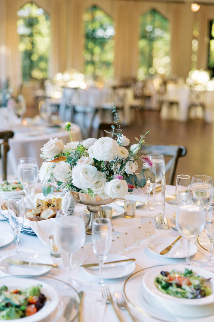 Wedding reception table set with white floral centerpiece in a gold compote, crystal glassware, gold flatware, and place cards, photographed inside a sunlit historic estate reception space.