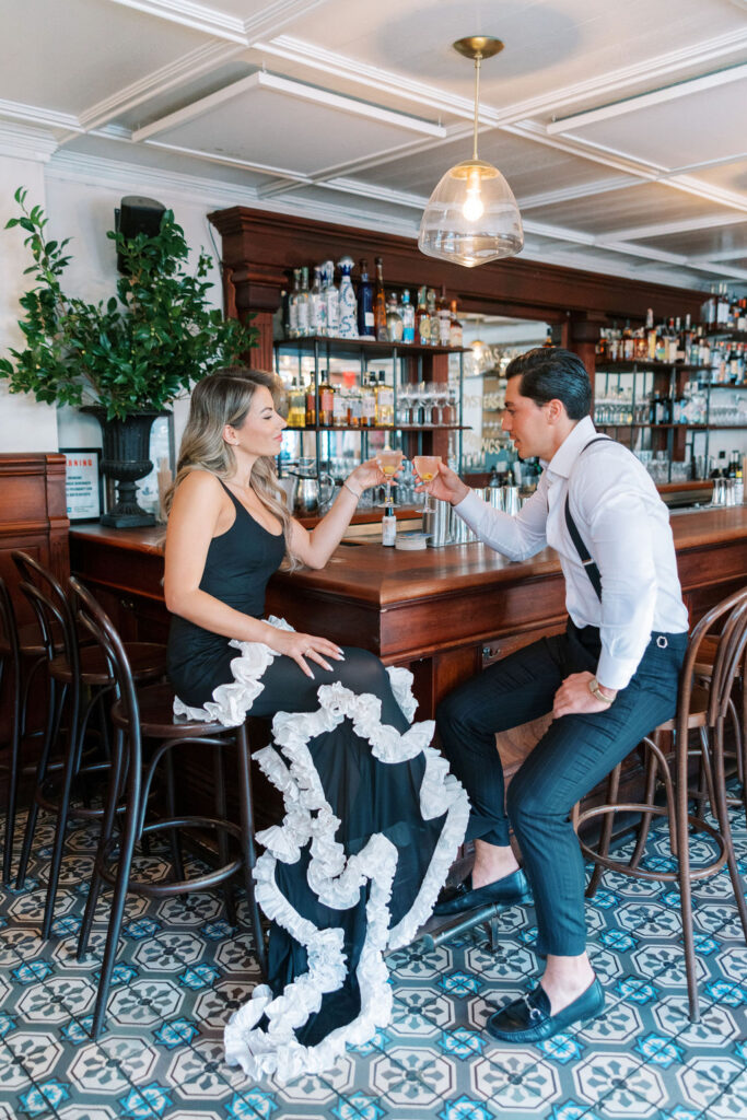 Couple sitting behind cafe bar in the West Village clink glasses posing for signature NYC wedding and engagement photography by Lauren Bliss, capturing timeless love stories