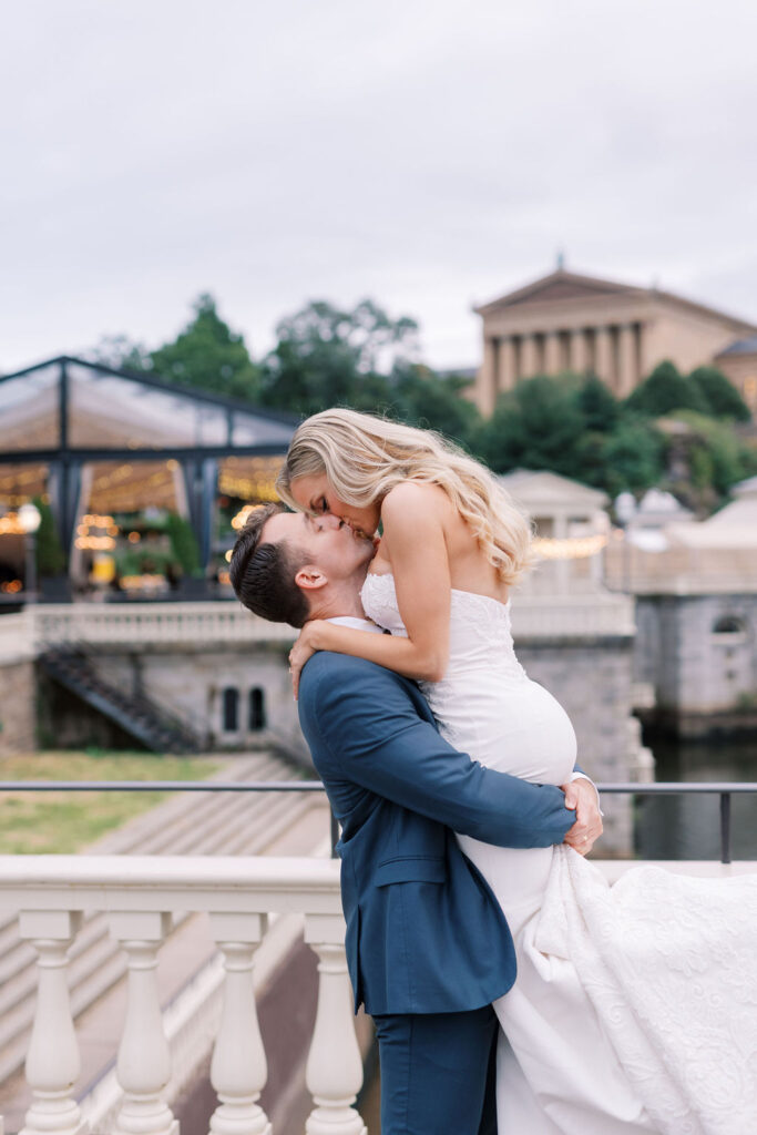 Groom lifts his bride and kisses her in front of a backdrop of the Philadelphia Art Museum and the Water Works Cescaphe venue