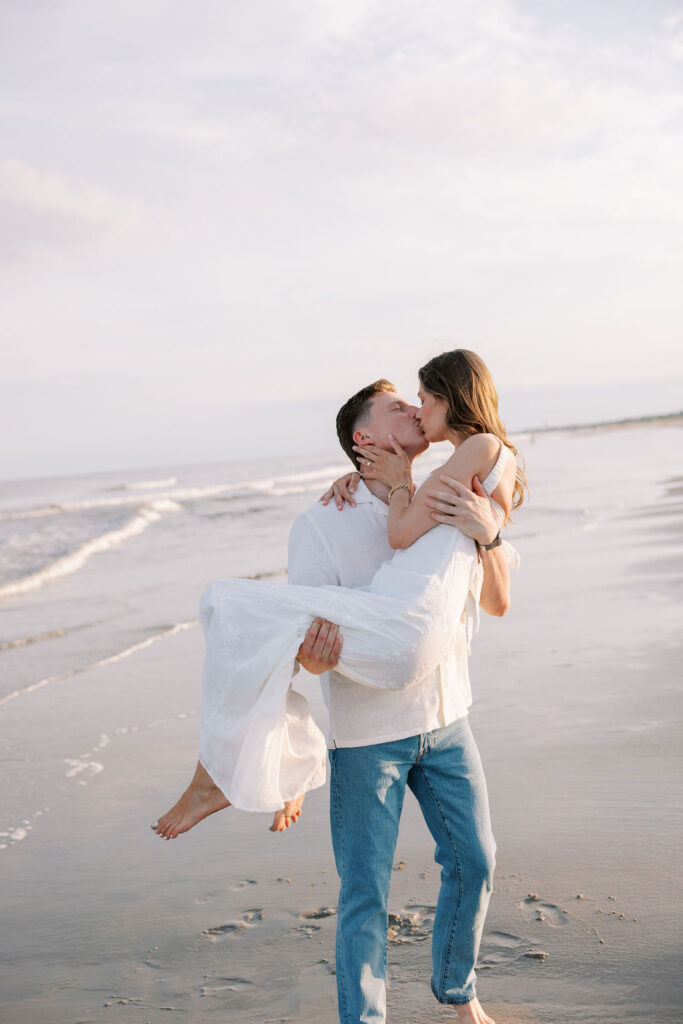 Man carries woman in his arms on the beach as they kiss during golden hour engagement portraits at Ocean City, NJ beach taken by Lauren Bliss Photography, full of warmth and romance