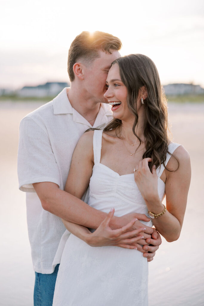 Man leans in to whisper in his fiance's ear during golden hour engagement portraits at Ocean City, NJ beach taken by Lauren Bliss Photography, full of warmth and romance