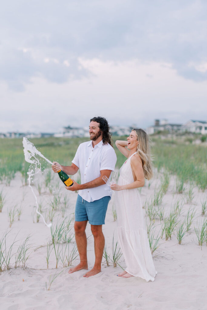Man pops a bottle of Veuve Clicquot champagne as woman laughs during elegant OCNJ beach engagement photoshoot by Lauren Bliss Photography, blending coastal beauty and timeless romance.