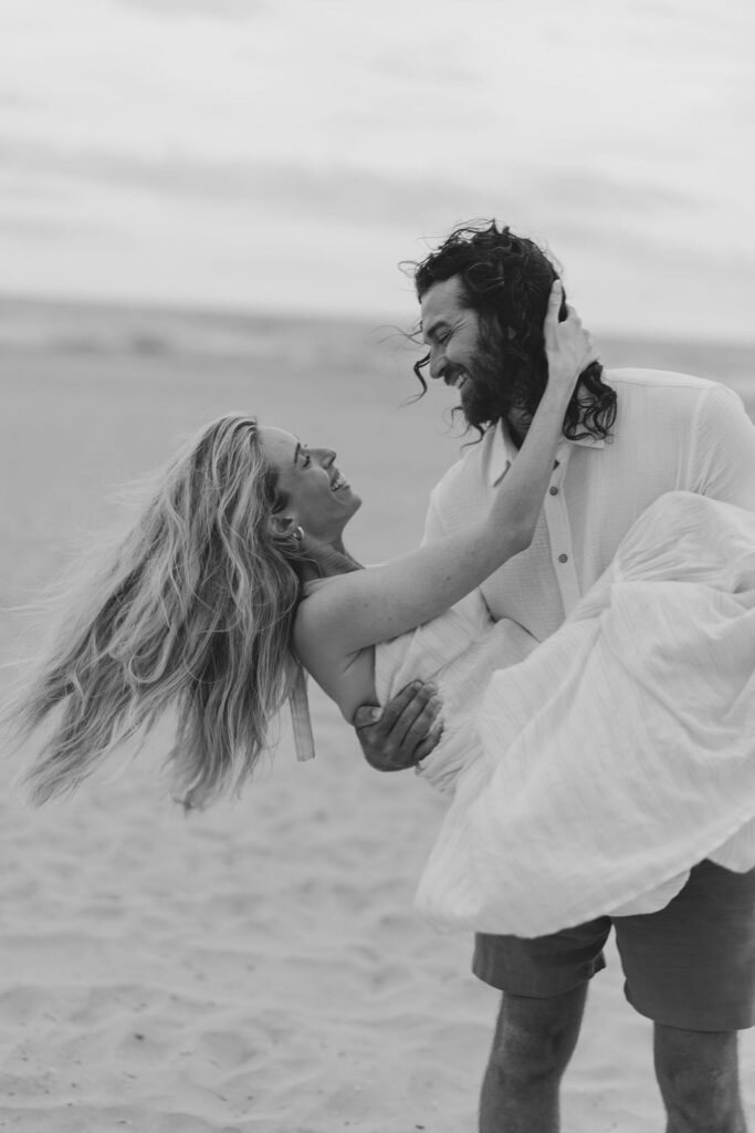 Man holds woman in his arms on the beach and dips her backwards during candid and joyful Ocean City, NJ beach engagement session captured by Lauren Bliss Photography