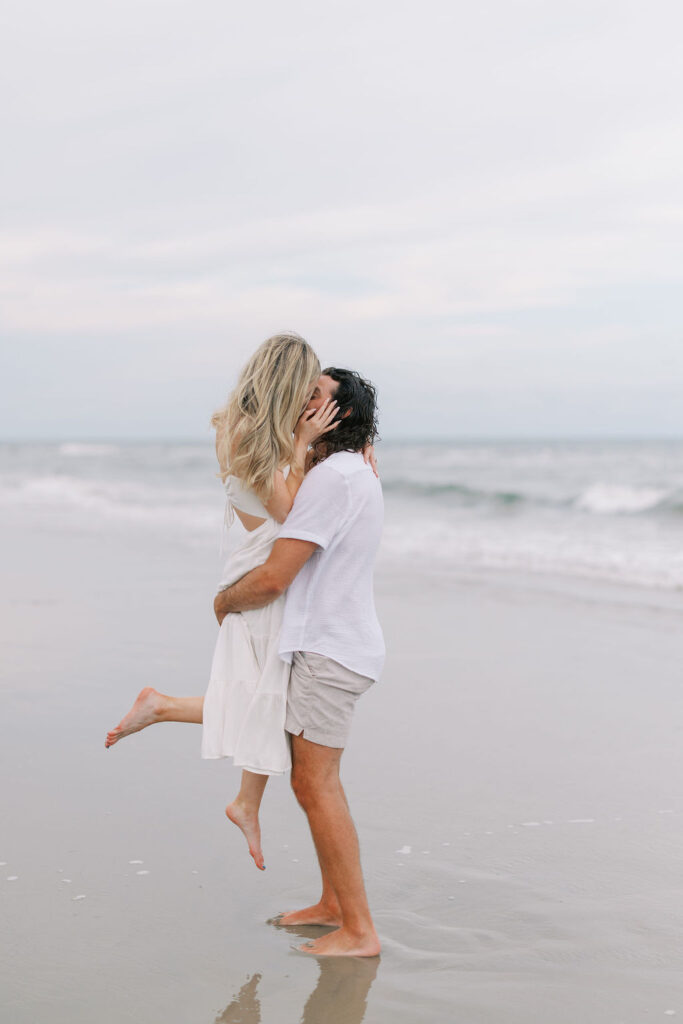 Man in casual outfit lifts woman in a white flowy dress during a dreamy Ocean City, NJ beach couple’s engagement session photographed by Lauren Bliss Photography.