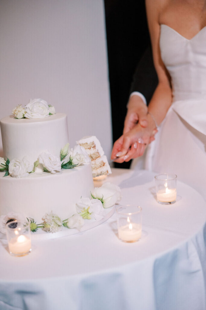 Bride and groom cutting a two-tier white wedding cake decorated with white roses at Tribeca Rooftop in New York City.
