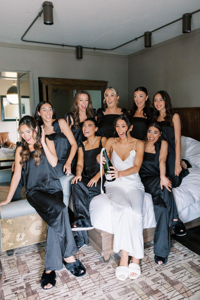 Bride pops champagne with bridesmaids in matching black pajamas before her Tribeca 360 rooftop wedding, photographed by NYC wedding photographer Lauren Bliss Photo.