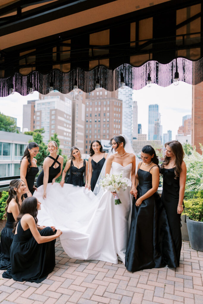 Bride surrounded by bridesmaids in chic black gowns, helping with her dress before rooftop vows at Tribeca 360 in Manhattan, photographed by Lauren Bliss Photo.