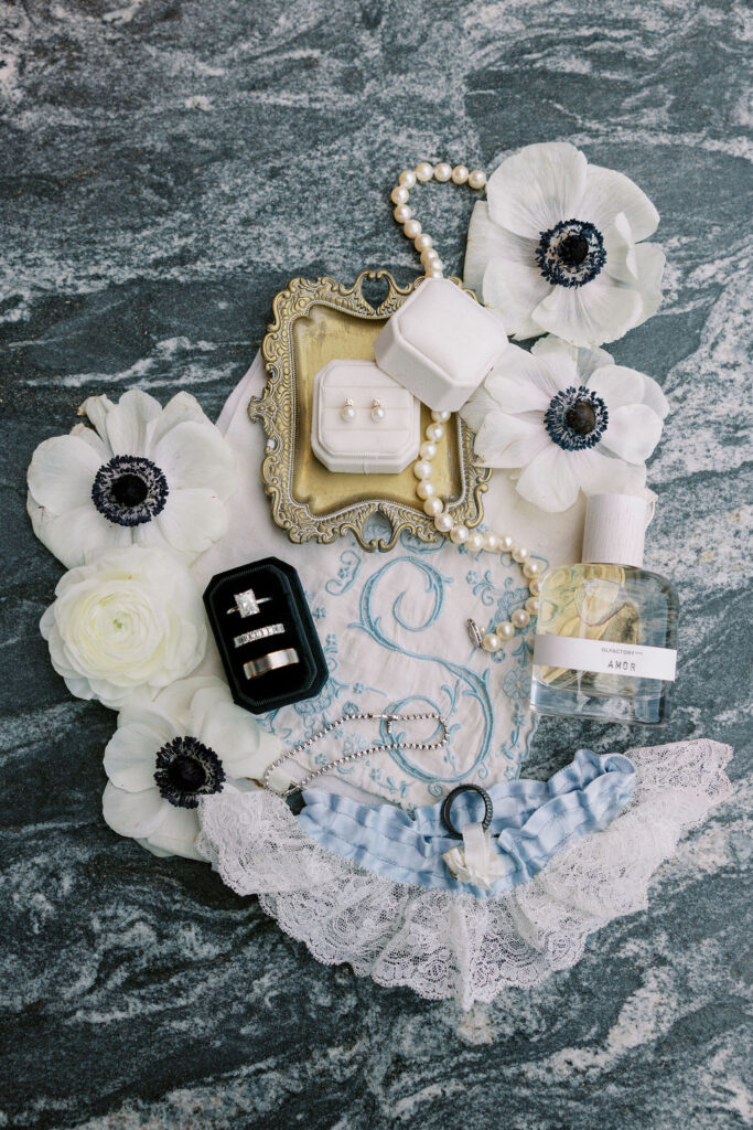 Elegant flat lay of bridal details with pearl earrings, wedding rings, lace garter, Amor perfume, and white florals at Tribeca Rooftop in New York City, captured by Lauren Bliss Photography.
