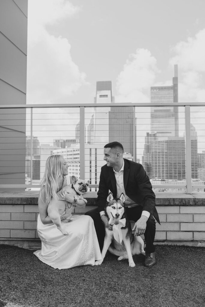 Black and white candid of Kelsey and Lee sitting on a rooftop with their Frenchie and Husky, sharing a playful engagement moment