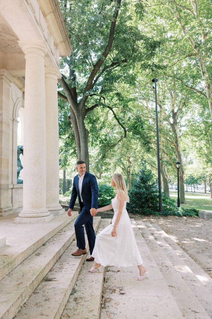 Kelsey and Lee walking hand-in-hand up the stone steps of the Rodin Museum, surrounded by tall trees and romantic architecture, photographed by Lauren Bliss Photography