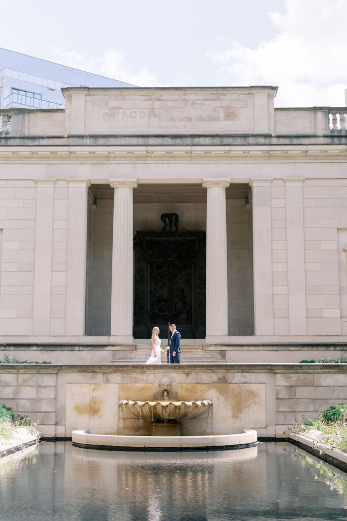 Kelsey and Lee holding hands in front of the Rodin Museum’s iconic gates and fountain during their Philadelphia engagement session