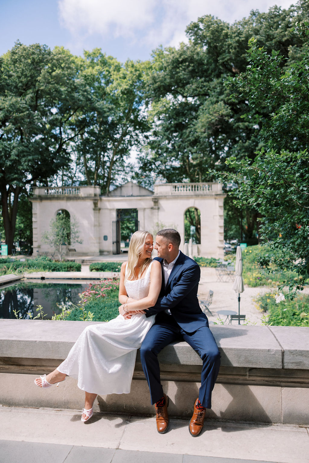 Kelsey and Lee share a joyful embrace while seated on a stone ledge near the reflecting pool at the Rodin Museum gardens in Philadelphia