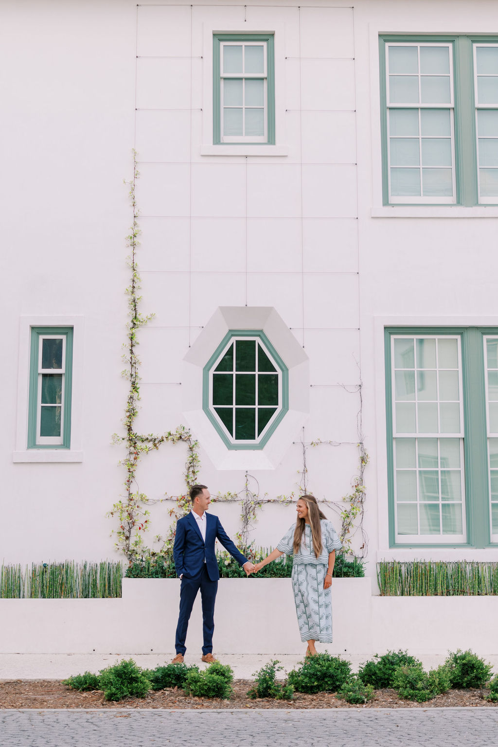 Couple holding hands and smiling while standing in front of a geometric window and white architectural wall in Alys Beach during their engagement session.