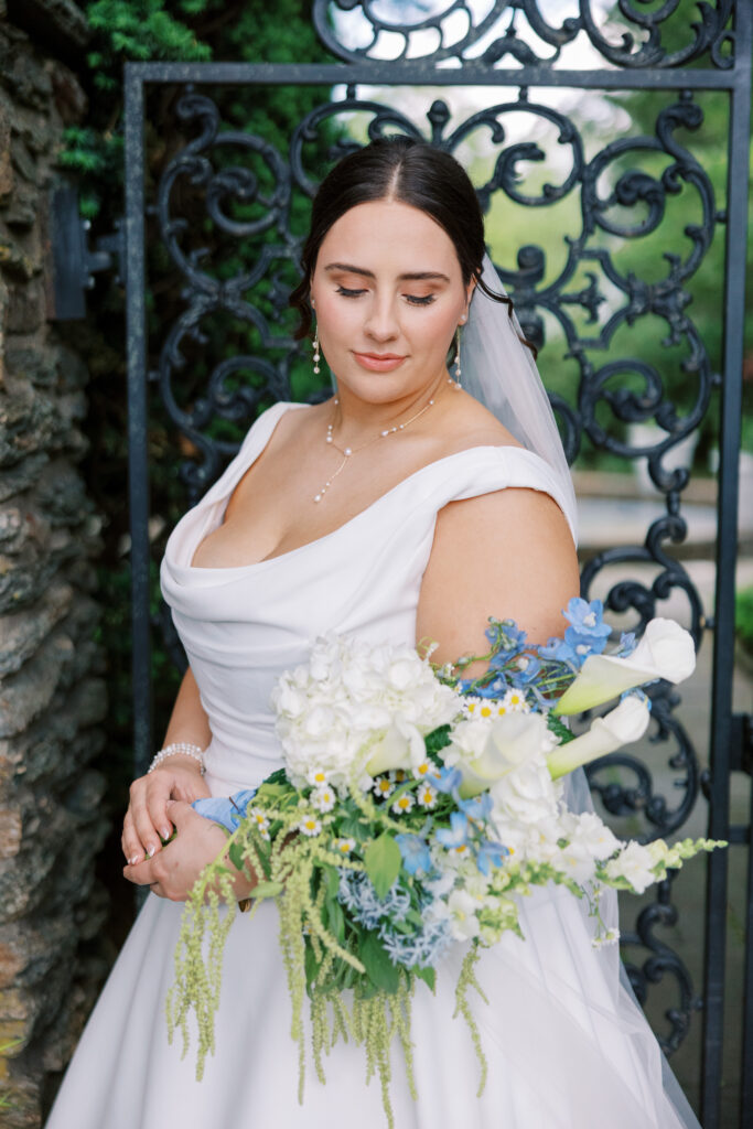 Bride posing in front of an ornate iron gate at Drumore Estate, holding a white and blue floral bouquet, featuring an off-the-shoulder wedding gown, veil, and garden estate backdrop.