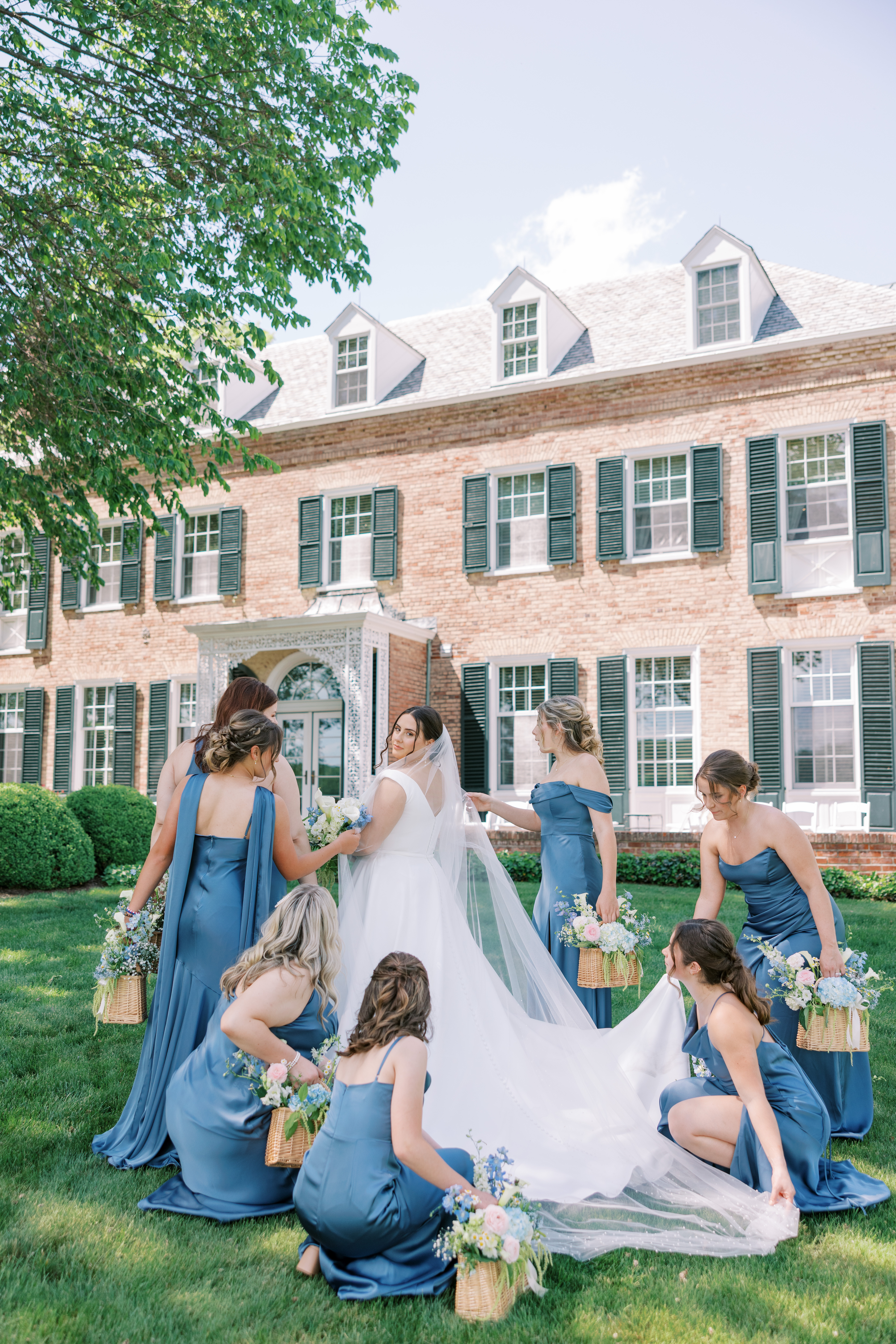 Bride is helped by six bridesmaids in satin blue dresses on the front lawn of the Drumore Estate wedding venue captured by Lauren Bliss Photography