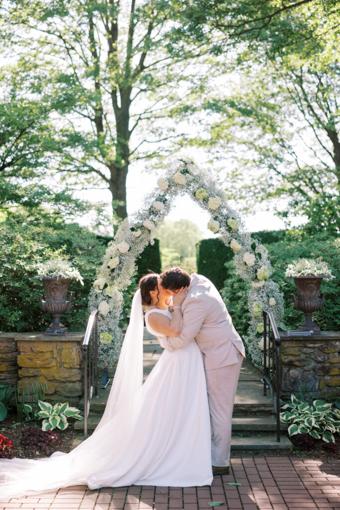 Bride and groom kissing beneath a white floral ceremony arch during an outdoor garden wedding at Drumore Estate, surrounded by stone steps, greenery, and soft natural light.