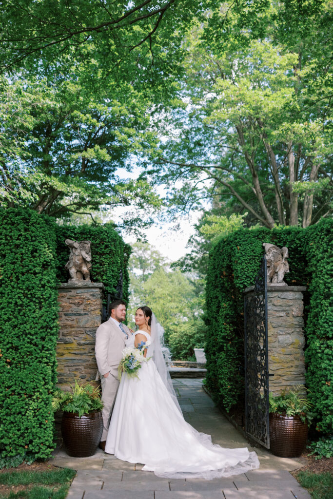 Bride and groom standing together at the stone and iron garden gate at Drumore Estate, surrounded by manicured greenery, featuring a flowing white wedding gown and light neutral groom attire.
