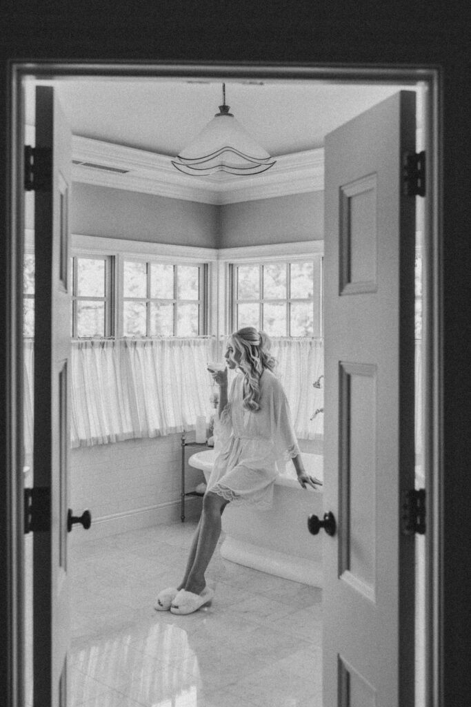 Black and white bridal prep portrait inside Pomme, featuring the bride in a robe sitting on a bathtub ledge with soft window light, captured by Lauren Bliss Photography.