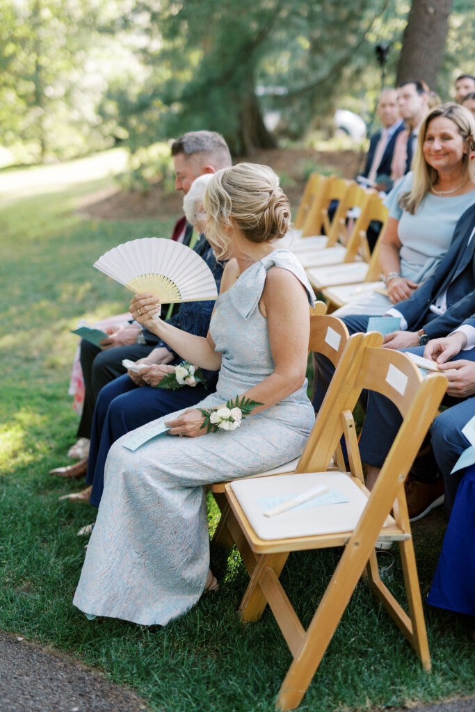 Wedding guests seated outdoors at Pomme in Radnor, Pennsylvania, with a mother of the bride using a hand fan on a warm summer wedding day, captured by Lauren Bliss Photography.