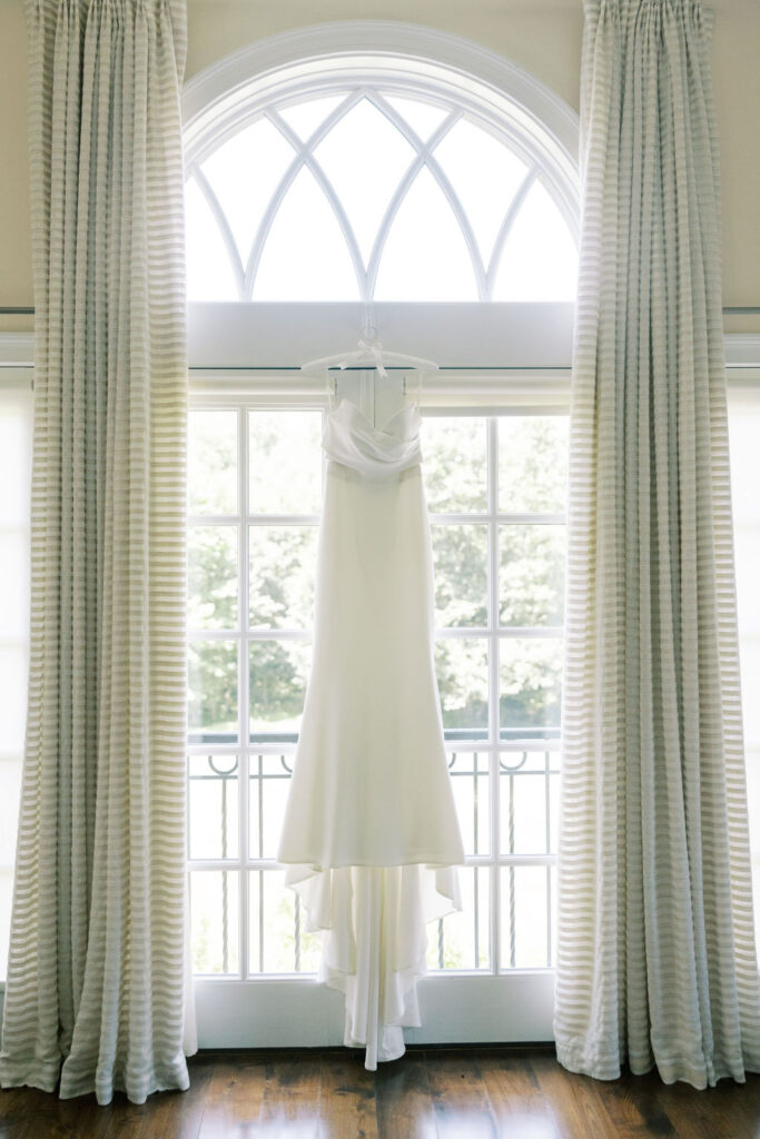 Bridal gown hanging in a bright window framed by soft curtains at Pomme in Radnor, photographed for elegant wedding detail inspiration by Lauren Bliss Photography.