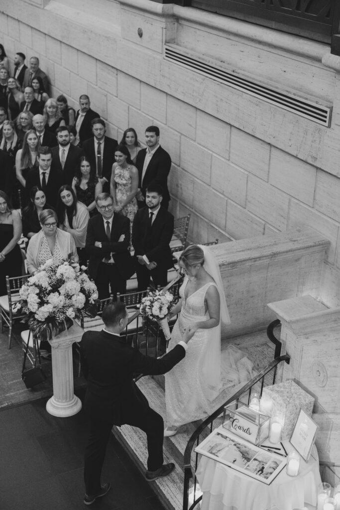 Black and white candid of a bride walking down the staircase at Union Trust while the groom reaches for her hand, surrounded by emotional wedding guests.