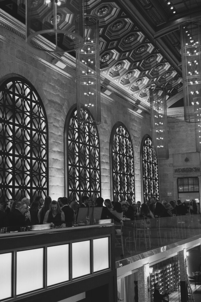 Black and white reception scene at Union Trust featuring its iconic arched windows, geometric ironwork, and dramatic hanging chandeliers during cocktail hour.