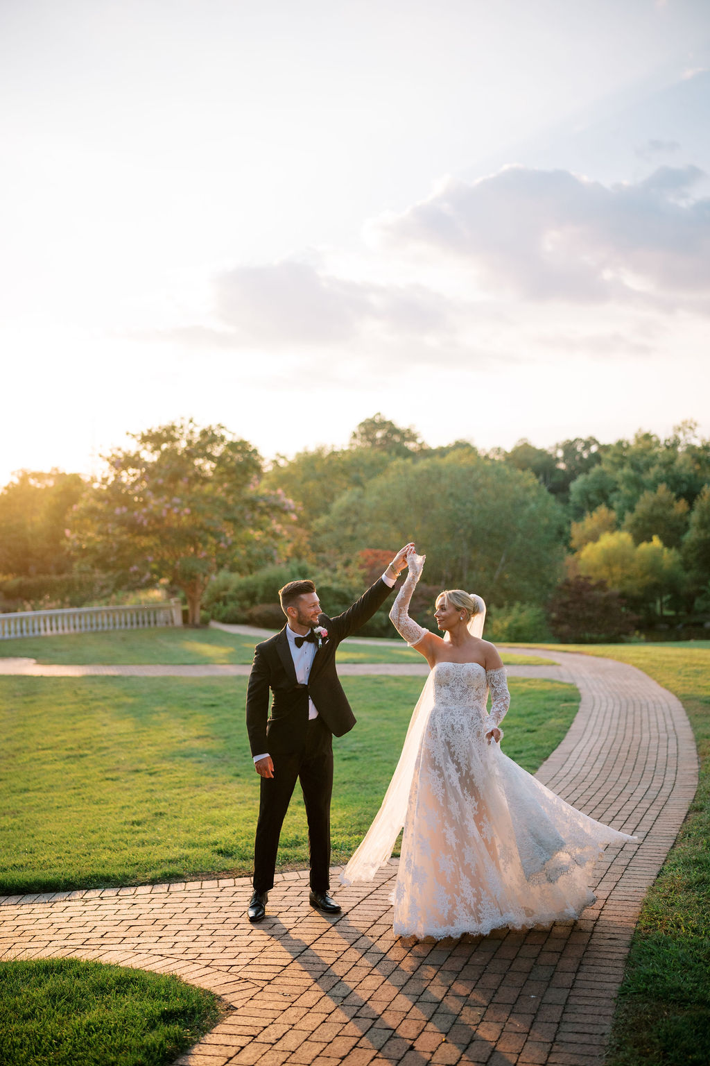 Groom twirls bride on a brick lined pathway with the sunset in the background during their wedding at Ashford Estate in NJ