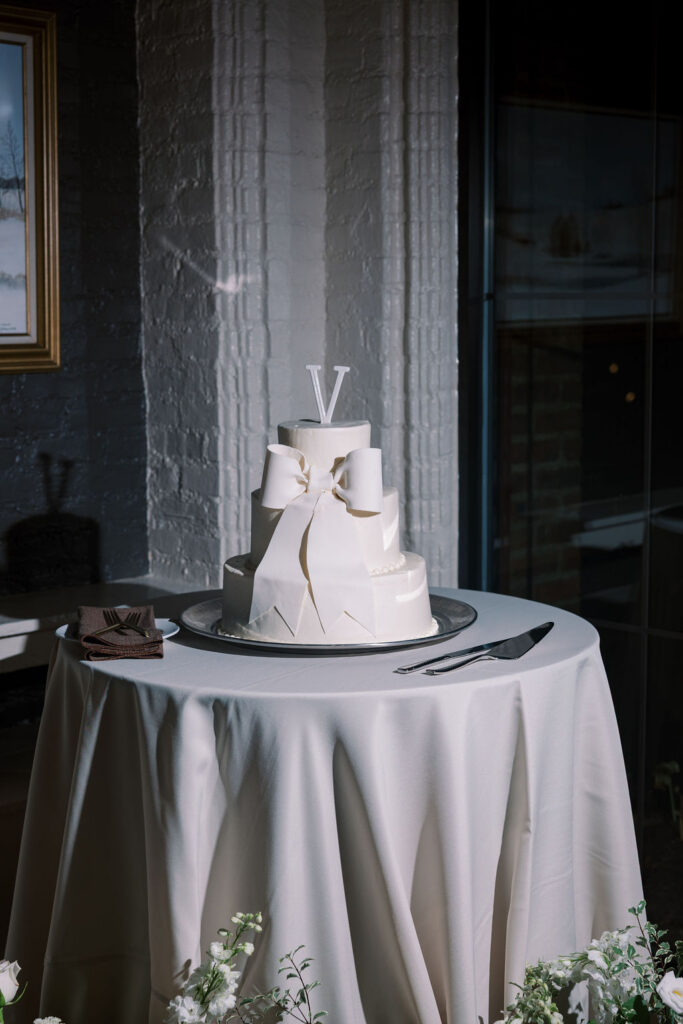 Three tiered white wedding cake with a large structural bow on the front and a simple monogram "V" cake topper