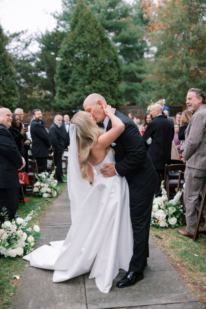 Bride and groom stop for a dramatic kiss at the end of the ceremony aisle during their outdoor wedding at Aldie Mansion