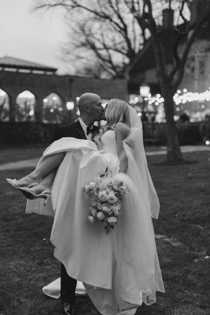 Black and white portrait of a groom carrying a bride with the Aldie Mansion's venue lights glowing in the background