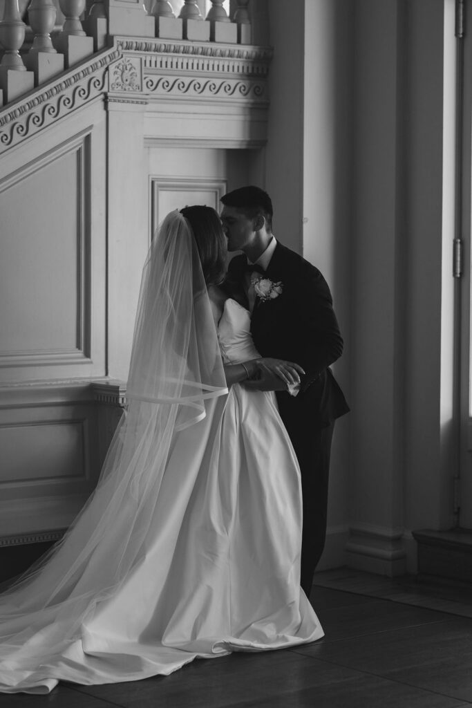 Black and white first kiss by the window of the grand staircase at a wedding at Cairnwood Estate