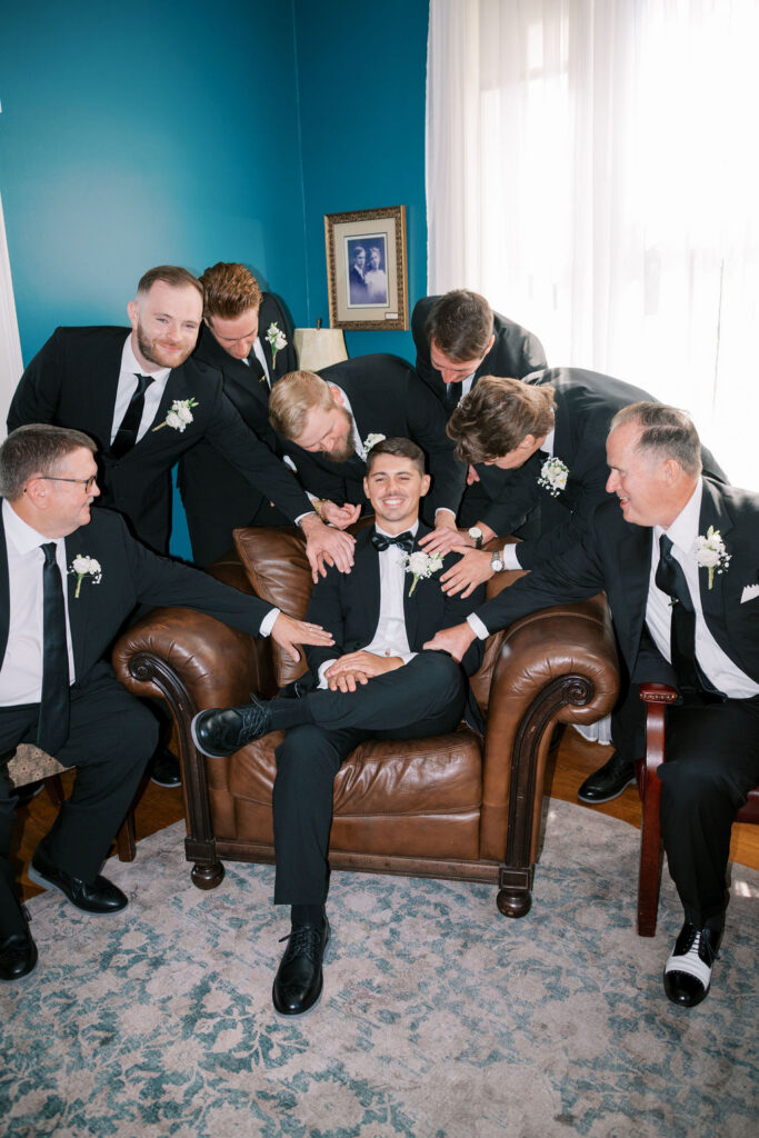 Groom and groomsmen in black tuxedos laughing in a teal getting-ready room