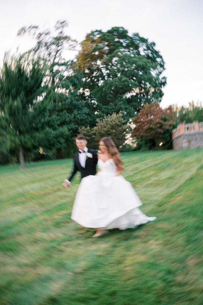 Motion-blur portrait of bride and groom running across the lawn at a wedding at Cairnwood Estate in Bryn Athyn, PA