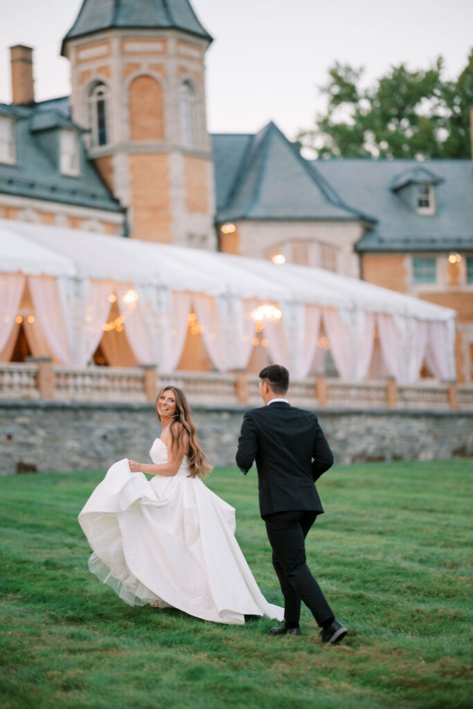 Bride and groom running toward the glowing terrace tent at dusk at a wedding at Cairnwood Estate, Bryn Athyn, PA