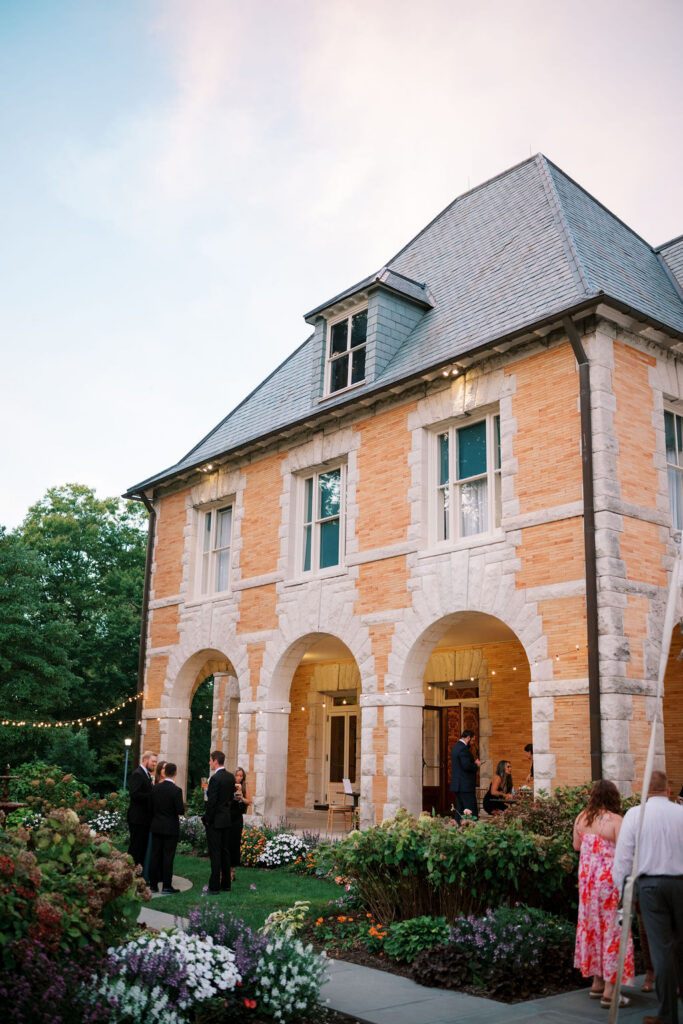 Guests at cocktail hour in the garden beneath the arched stone loggia at a wedding at Cairnwood Estate