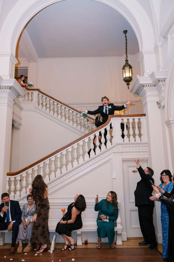 Groomsman tossing petals from the grand staircase as guests cheer at a wedding