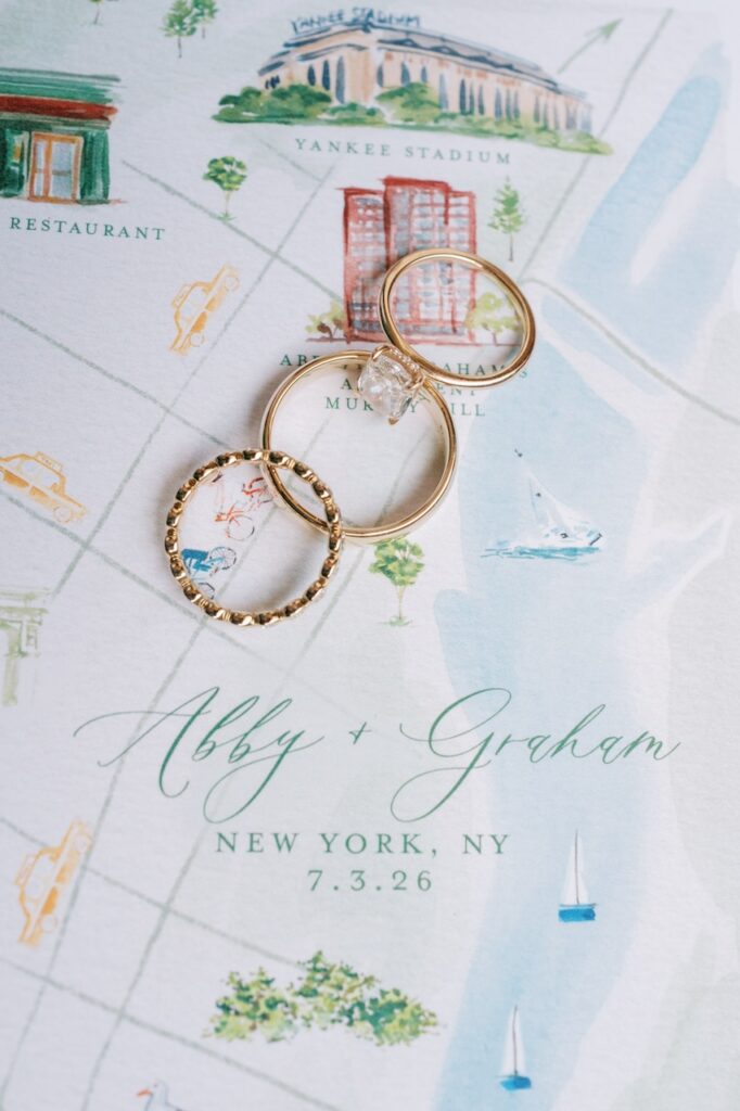 Three gold rings sit on top of a watercolor wedding invitation that reads 'Abby + Graham, New York, NY, 7.3.26'