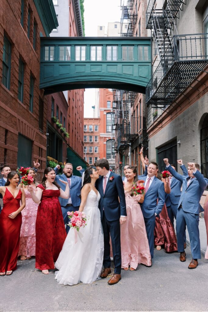 Bride and groom kiss surrounded by their wedding party under a green covered bridge in Tribeca NYC