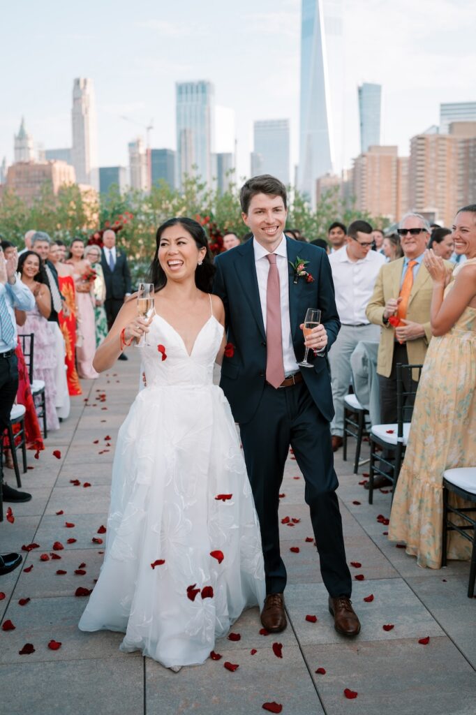 Tribeca Rooftop wedding photographer capture of couple holding champagne glasses during their ceremony recessional rose petal toss