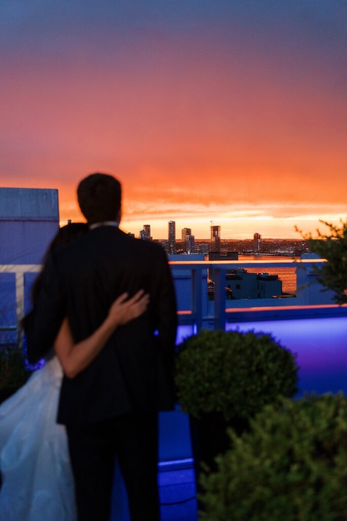 Bride and groom stand close together and watch the sunset over the Manhattan skyline from Tribeca Rooftop