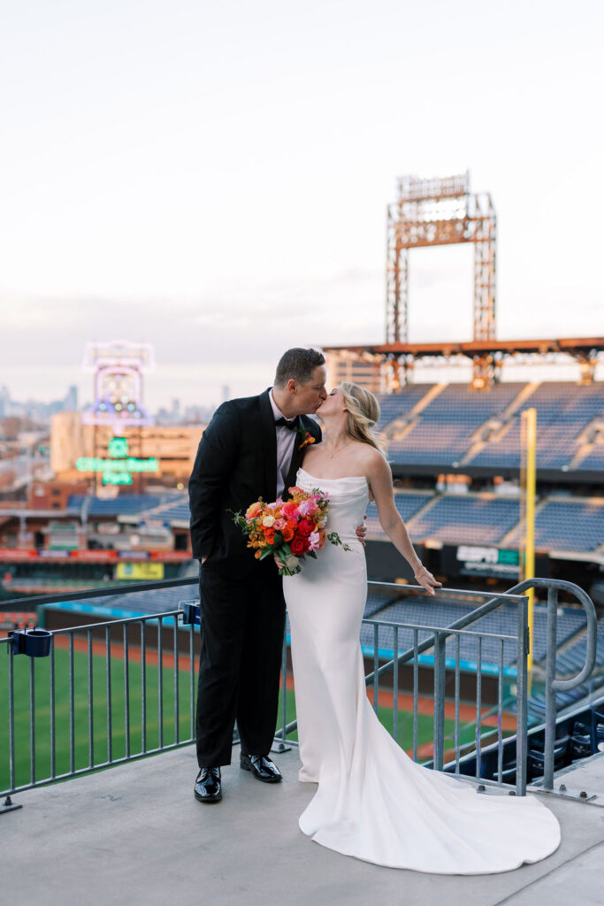 Kissing bride and groom with Citizens Bank Park sign blurred in the backdrop and glowing in front of the Philadelphia skyline