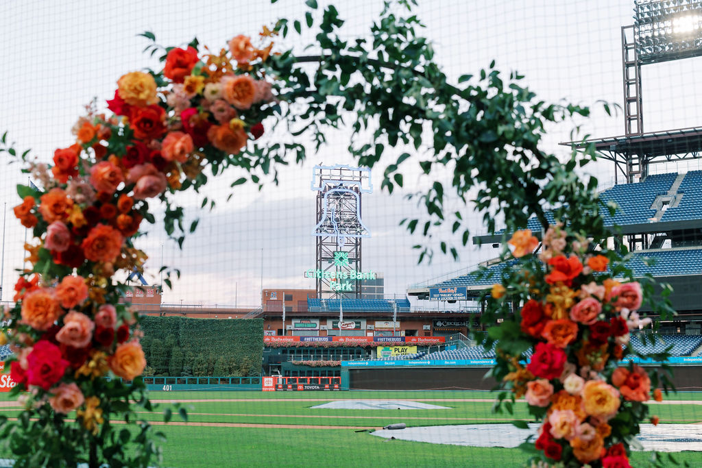 Orange, red, and peach floral ceremony arch frames the Citizens Bank Park neon sign in the background during a Phillies baseball wedding
