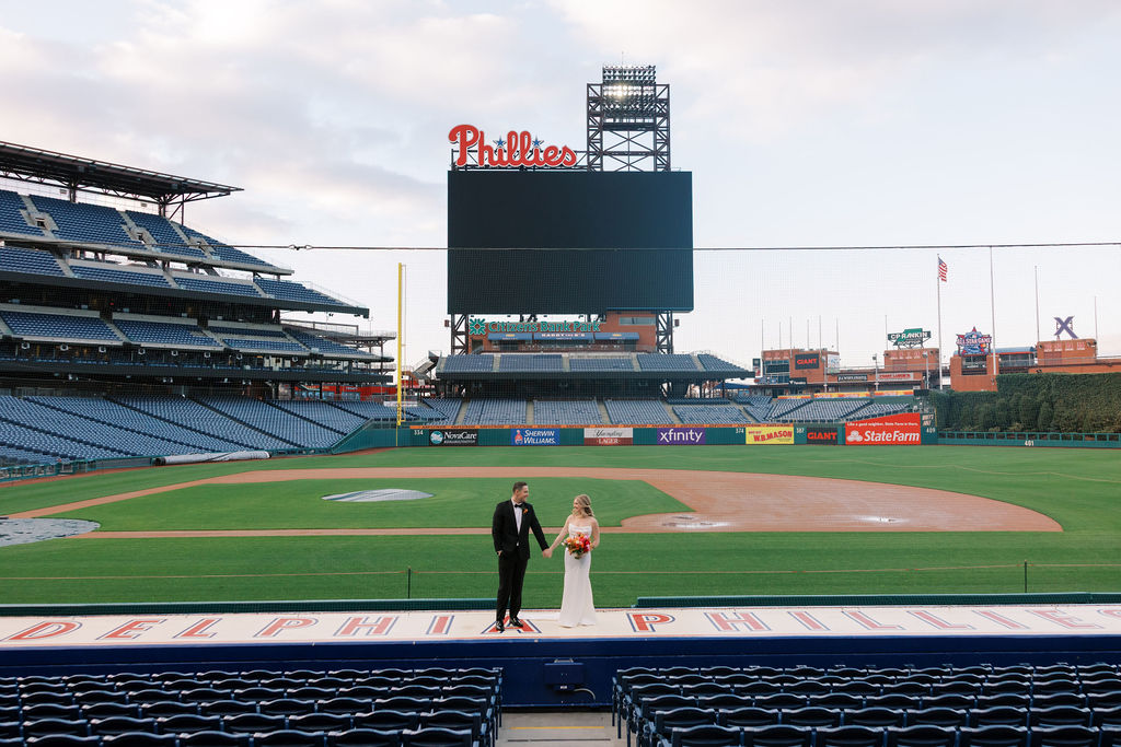 Bride and groom hold hands and stand on top of the dugout at Citizens Bank Park in front of the Phillies sign