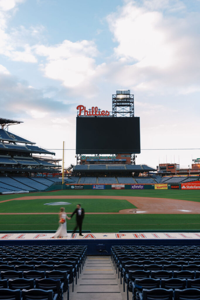 Bride and groom hold hands and walk across the dugout at Citizens Bank Park in front of the Phillies sign