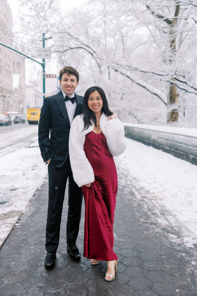 Couple poses in Central Park NYC with snow falling all around them and snow covered branches in the backdrop