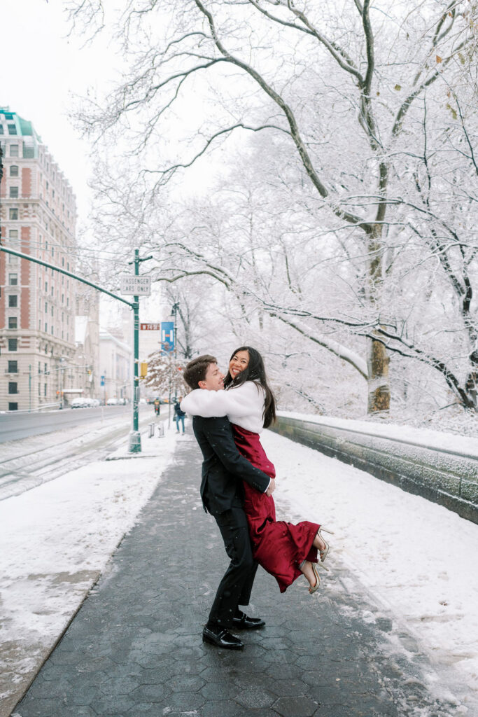 Man lifts a woman in a red silk dress and white fur wrap in Central Park with snow falling all around them during winter engagement in NYC