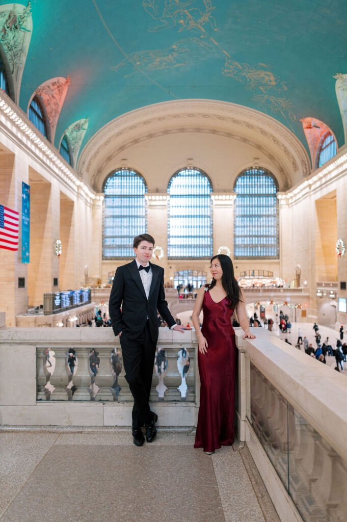 Couple in formalwear poses together on a landing inside Grand Central Terminal with large arched windows in the background during winter engagement in NYC