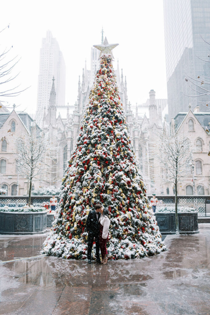Couple kisses in front of a tall red and gold decorated Christmas tree covered in snow