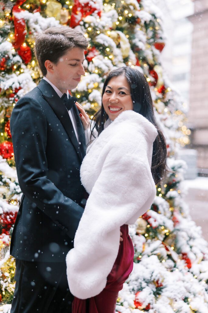 Man in a black tuxedo holds his fiance wearing a deep red silk dress and white fur wrap in front of a red and gold snow covered Christmas tree in NYC